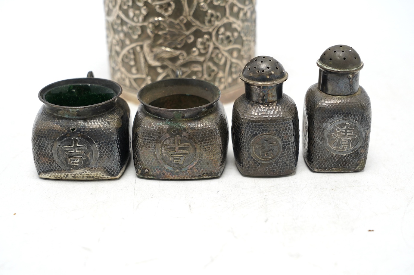 A Chinese embossed white metal cylindrical jar and cover, 11.2cm, and a similar white metal four piece cruet set. Condition - poor to fair